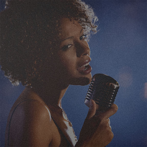 A photo of a black women who is holding a mike. The tone is dark blue with low saturation and grainy texture over it.